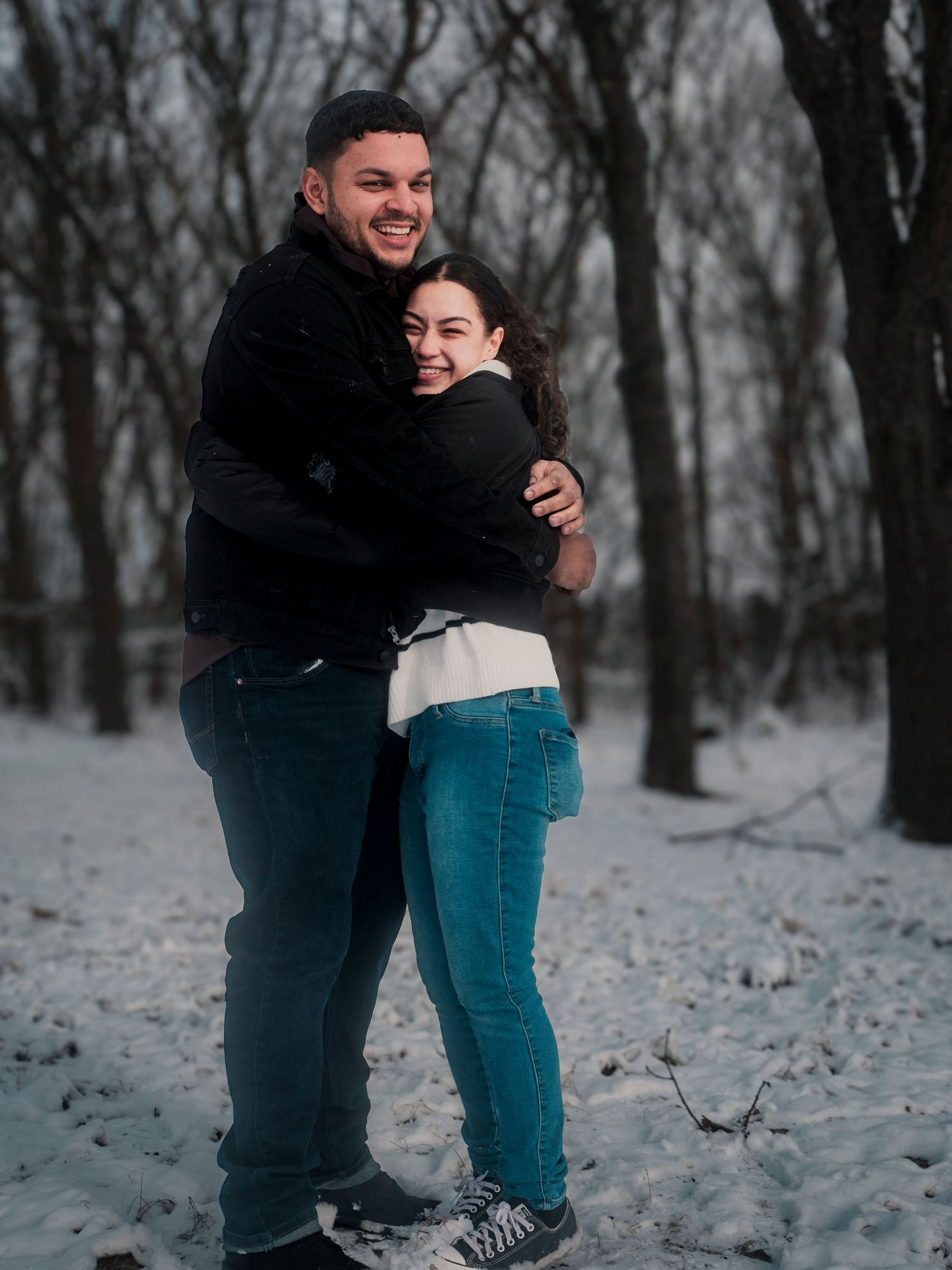 A couple shares an embrace while standing in a snowy winter forest landscape.