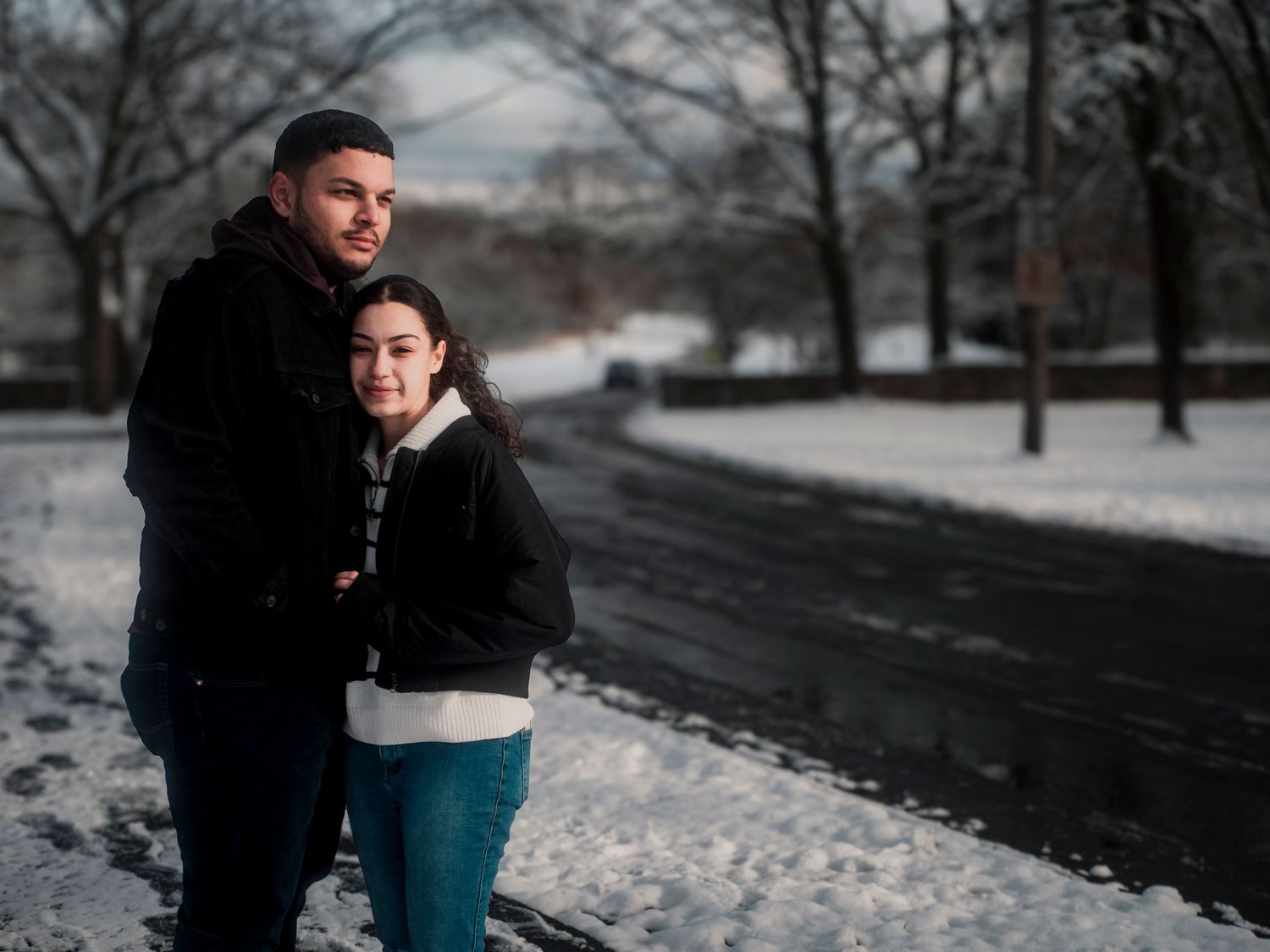 A couple shares a romantic moment in a snowy winter park setting, surrounded by bare trees and white ground.