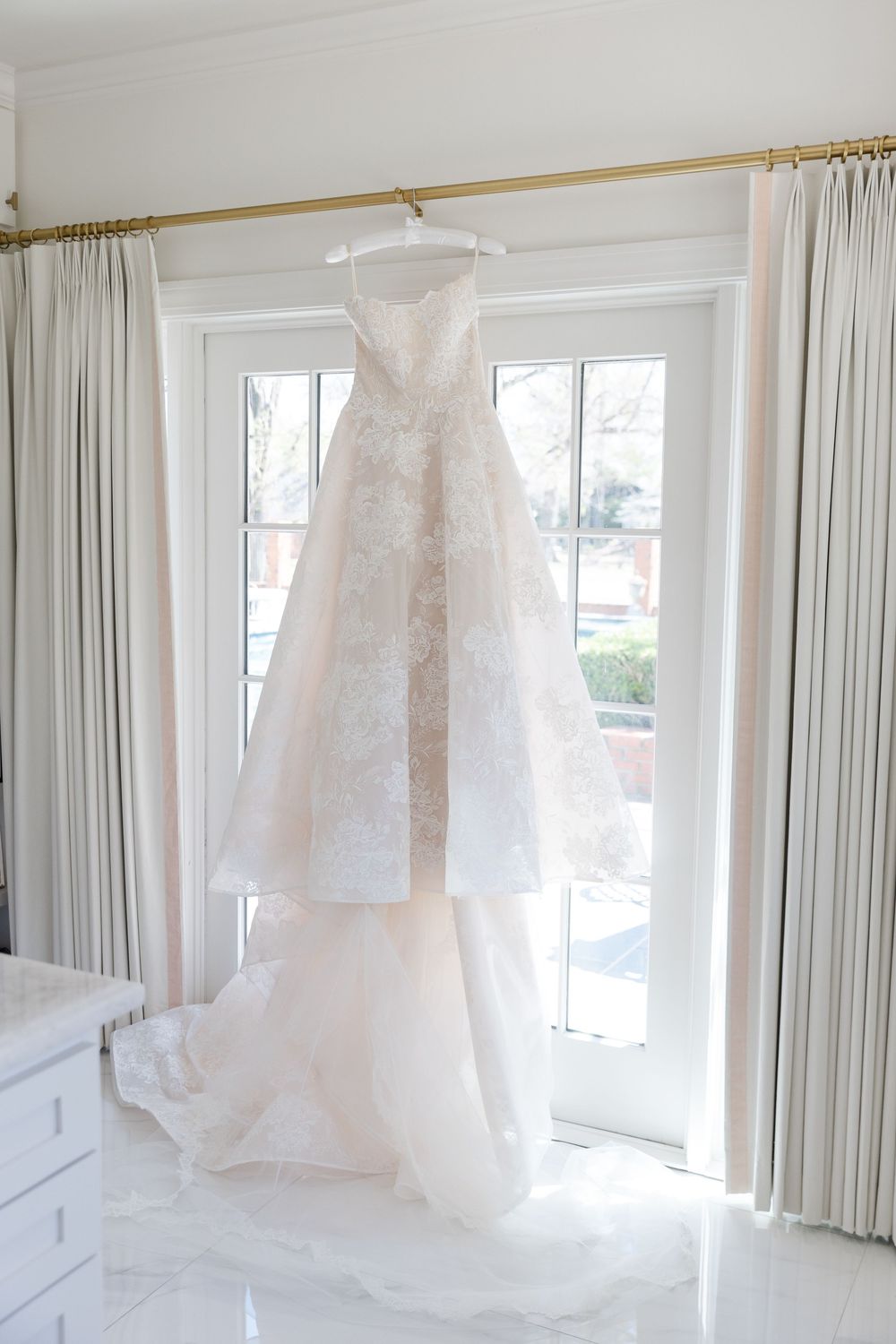 Elegant strapless lace A-line wedding gown hanging on a gold rod in front of white French doors.