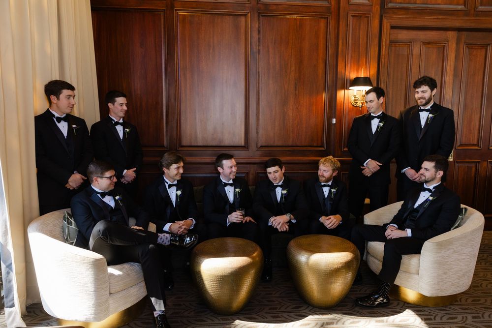 Groomsmen in black tuxedos gathered in an elegant wood-paneled room with gold accent furniture.