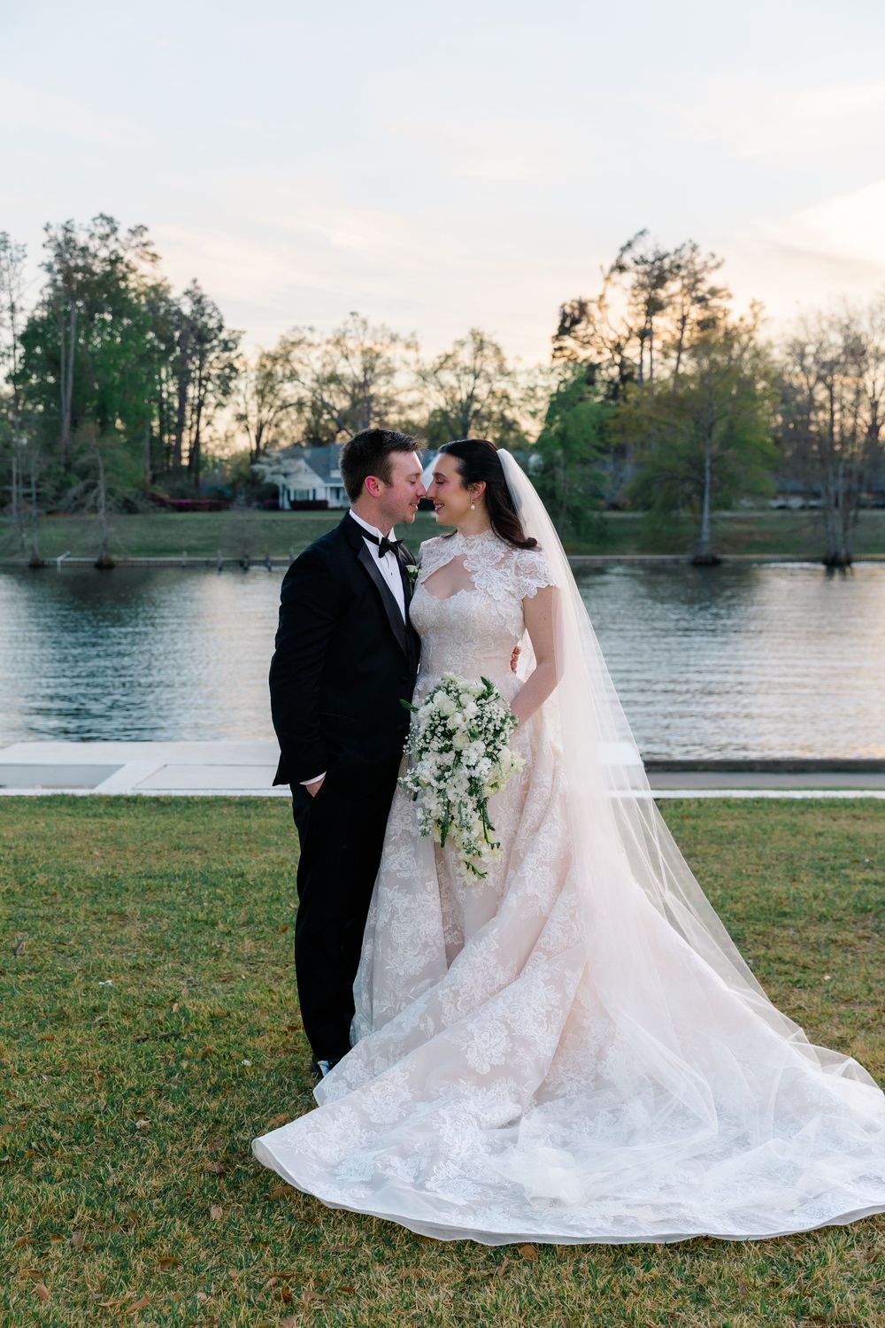 Bride and groom share a romantic moment by a lakeside at sunset, bride in lace gown.