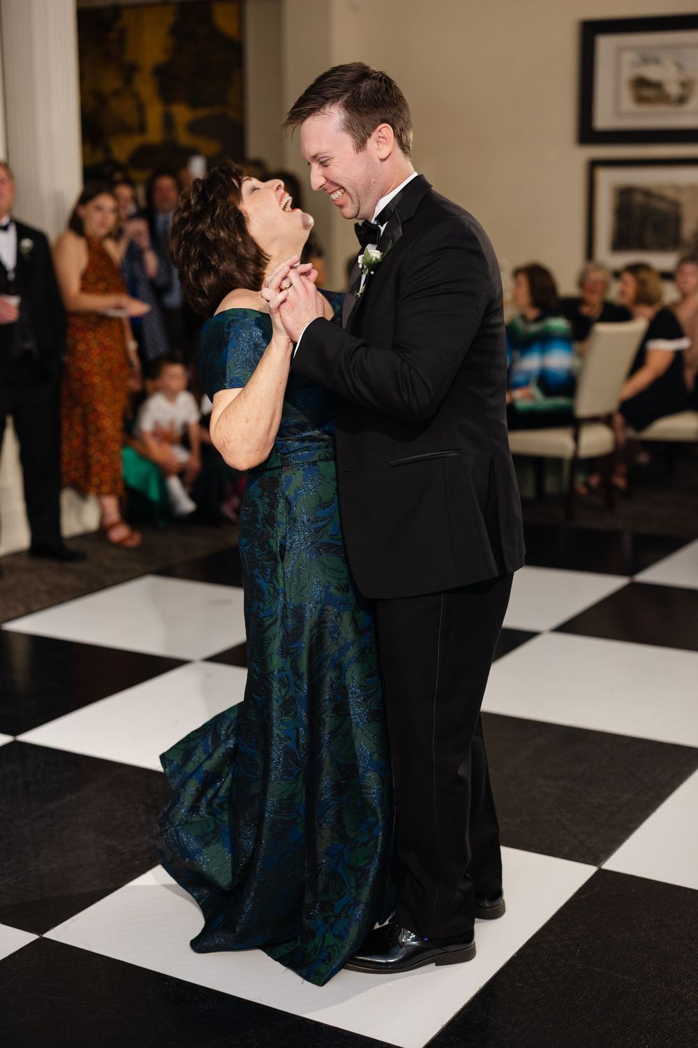 Groom in black tuxedo shares a joyful mother-son dance on a black and white checkered floor.