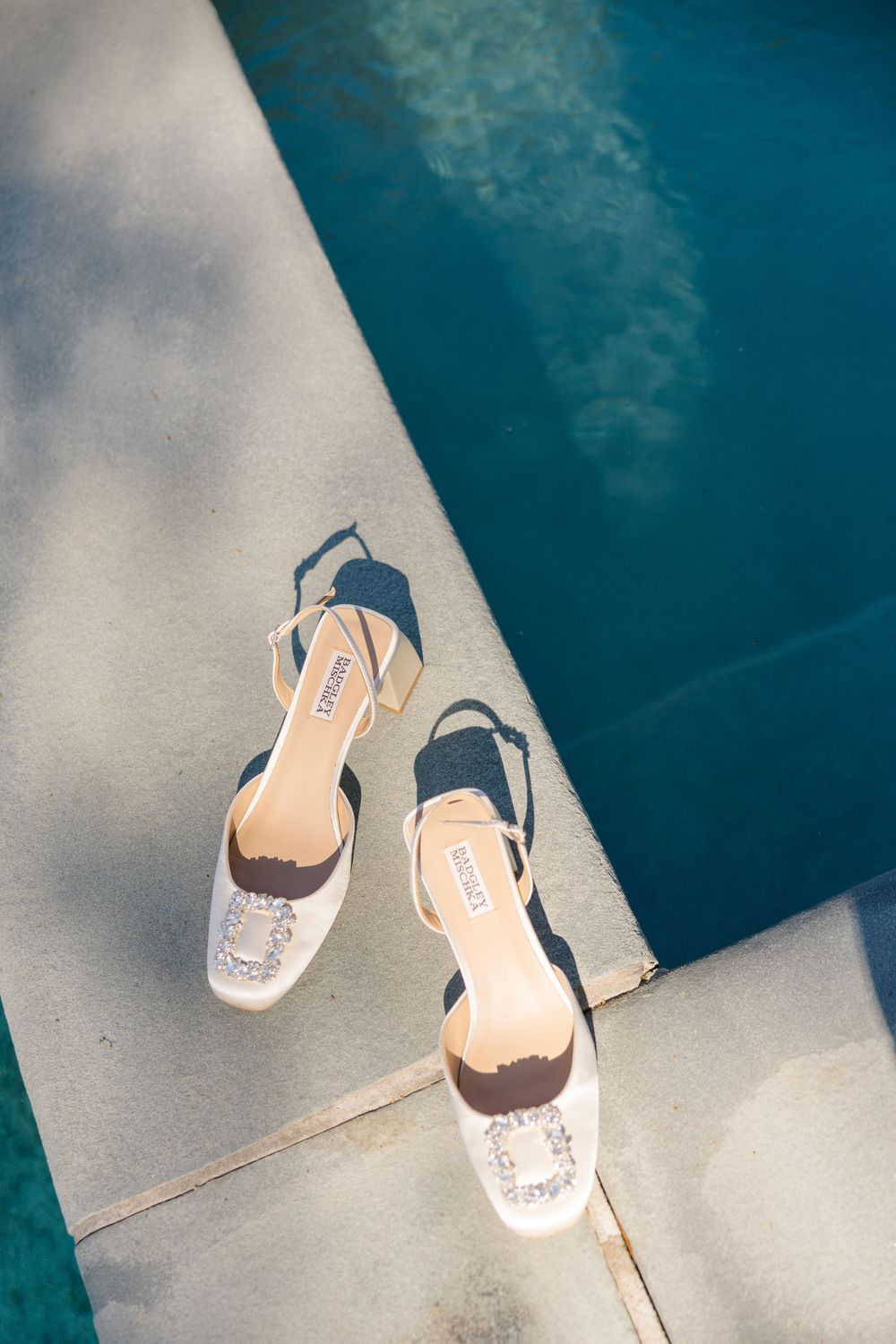 White bridal heels with crystal buckles placed on pool edge beside sparkling blue water.
