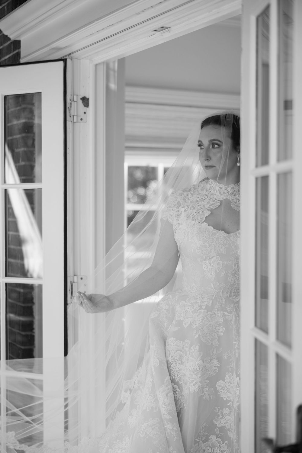 Black and white portrait of bride in lace gown and veil standing in white French doorway.