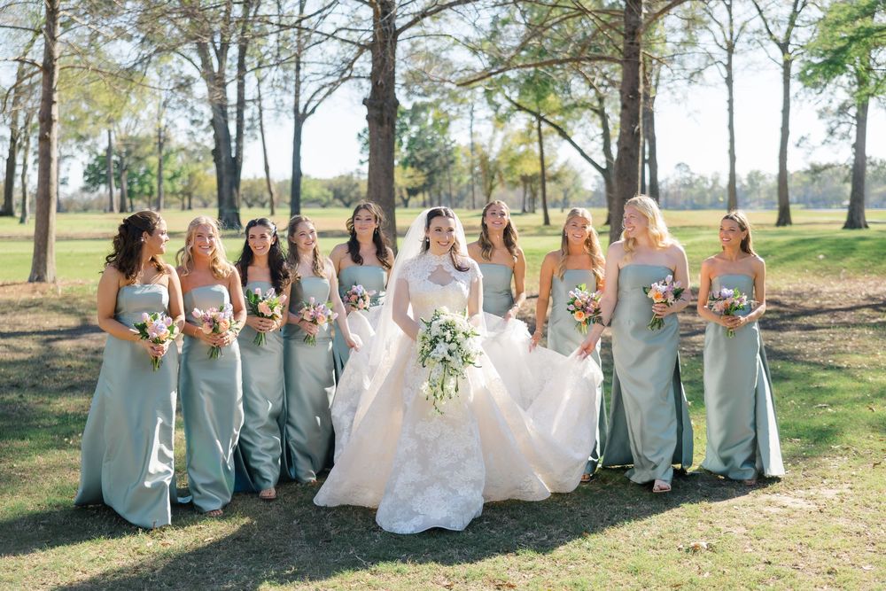 Bride in white ballgown poses with ten bridesmaids in sage green dresses holding colorful bouquets outdoors.