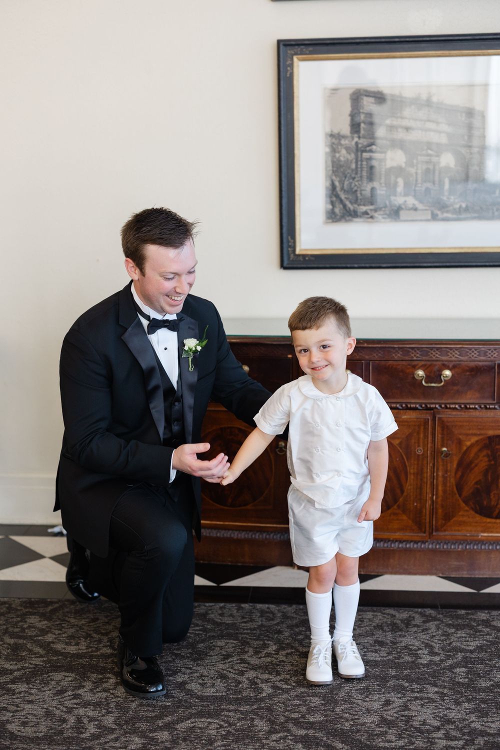 Smiling groom in black tuxedo kneels beside young ring bearer in white outfit indoors.