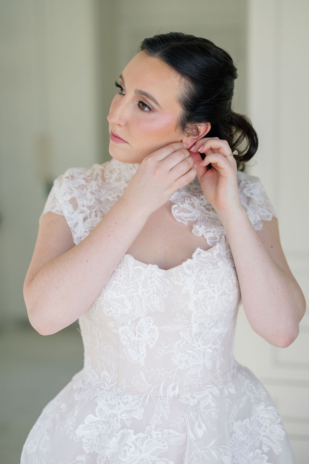 Dark-haired bride in lace wedding dress puts on earrings while getting ready before ceremony.