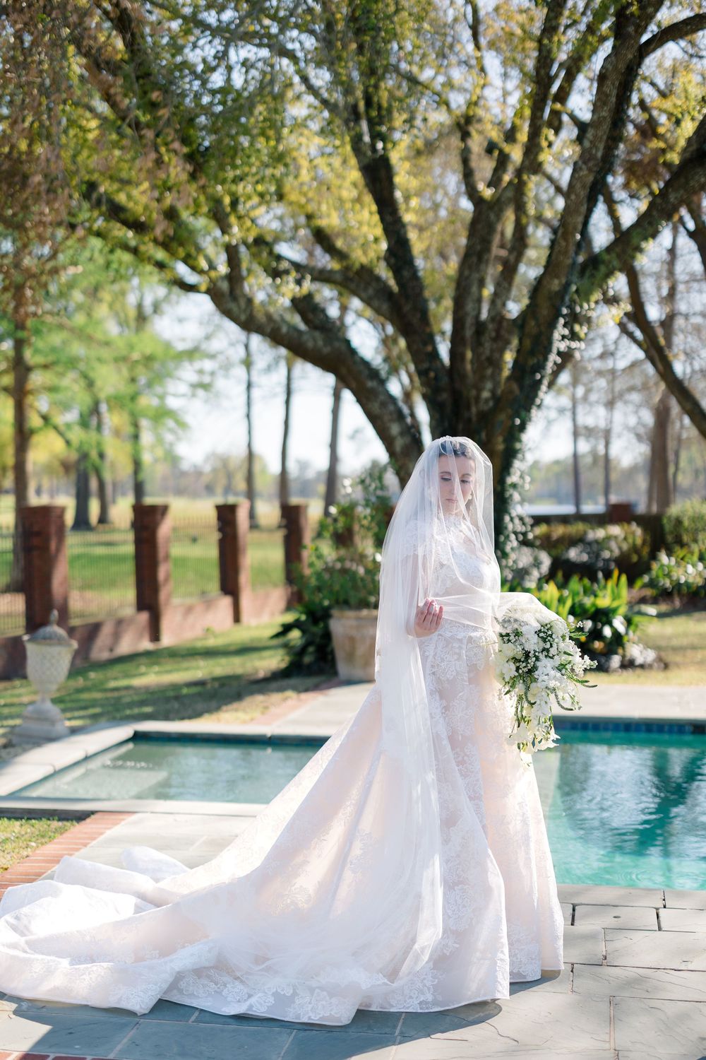 Bride in elegant lace wedding gown with long veil holding white bouquet beside a garden pool.