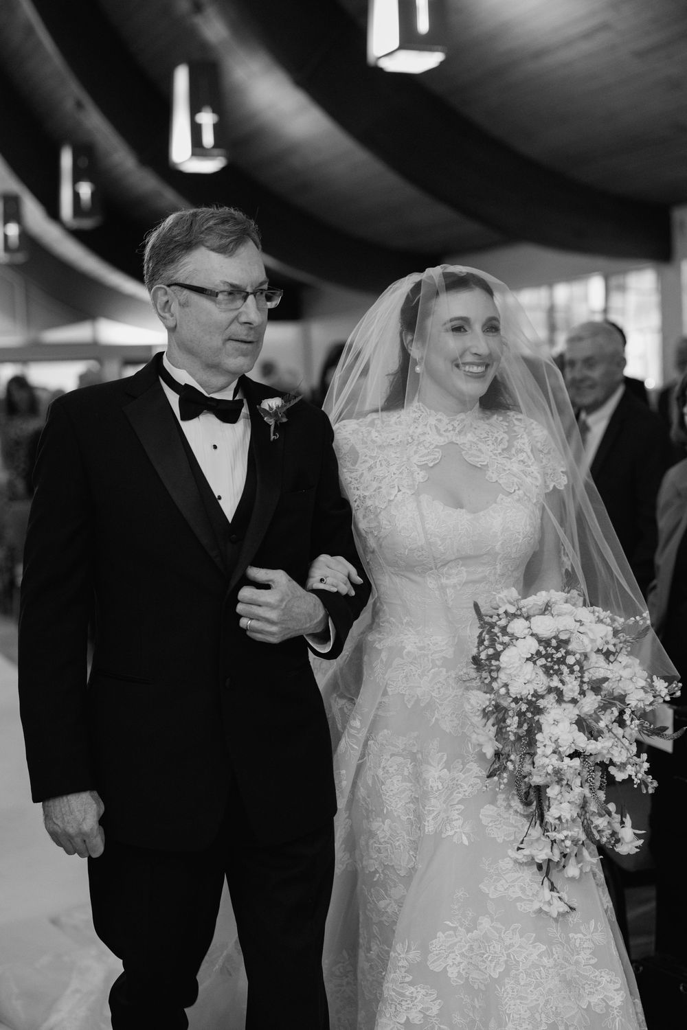 Black and white photo of smiling bride in lace gown and veil walking down aisle with father.