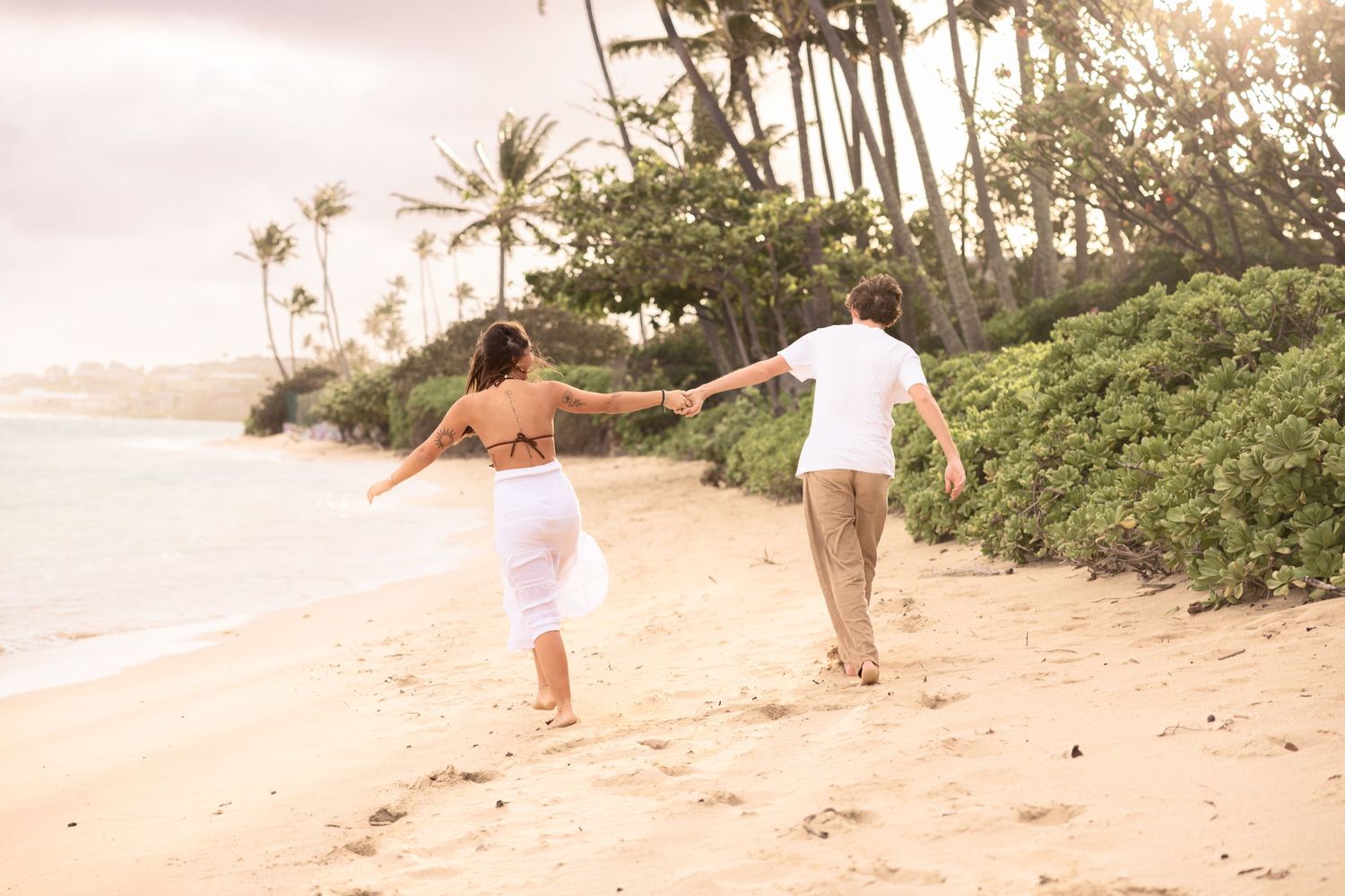 A couple walks along a sandy beach with palm trees and ocean waves in Hawaii during sunset.