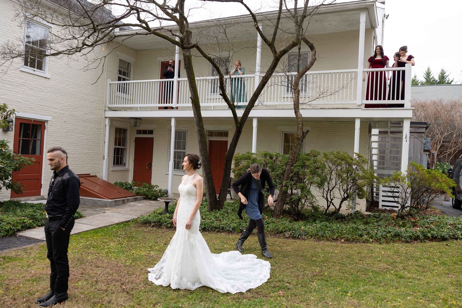 Wedding party members walk in formation across a grassy lawn outside a white colonial style building.