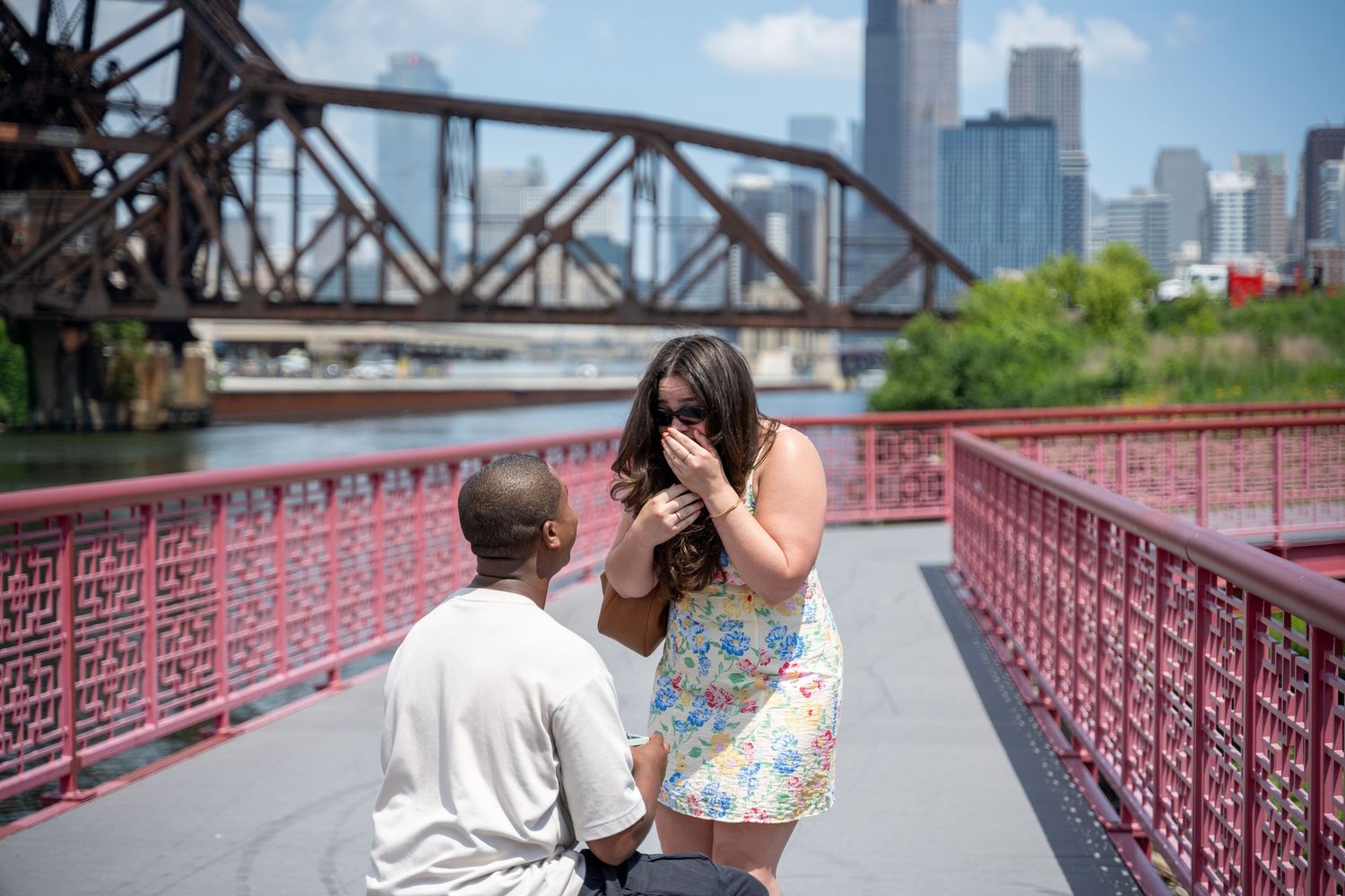 People enjoy views of the city skyline from a red-painted bridge walkway on a sunny summer day.