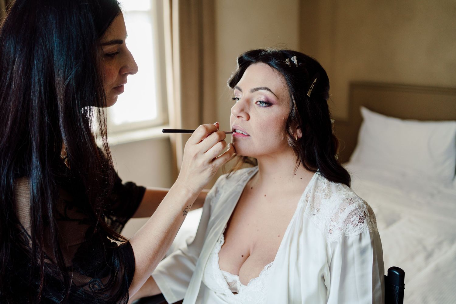 Professional makeup artist applying lipstick to bride in white robe during wedding morning preparation.