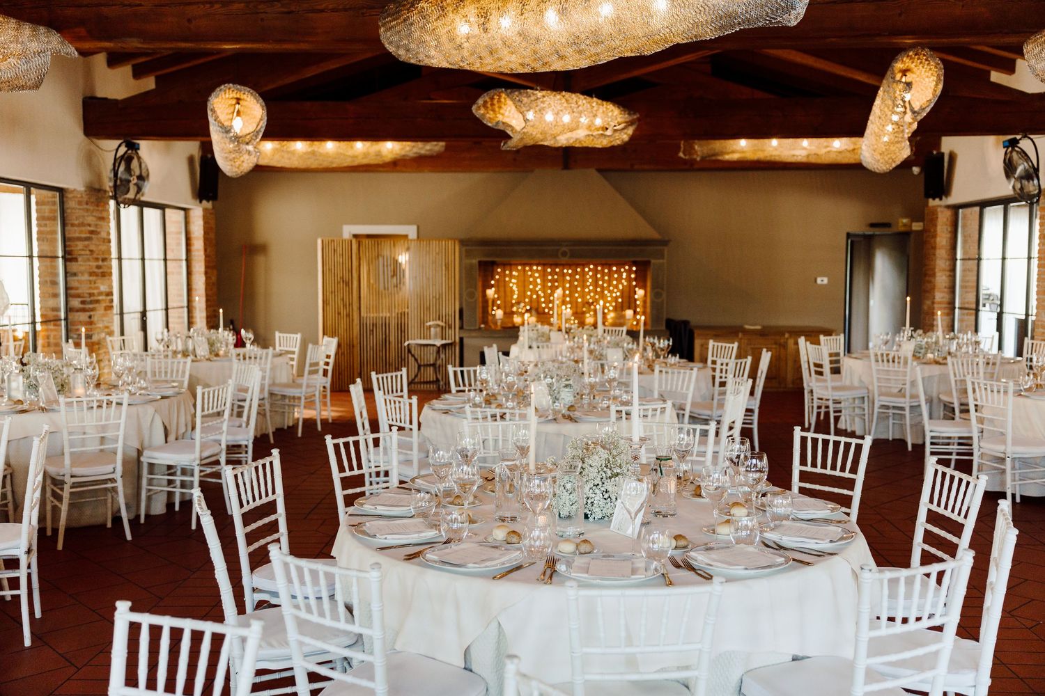 Bright wedding reception hall with round tables, white chiavari chairs, and decorative hanging lights.