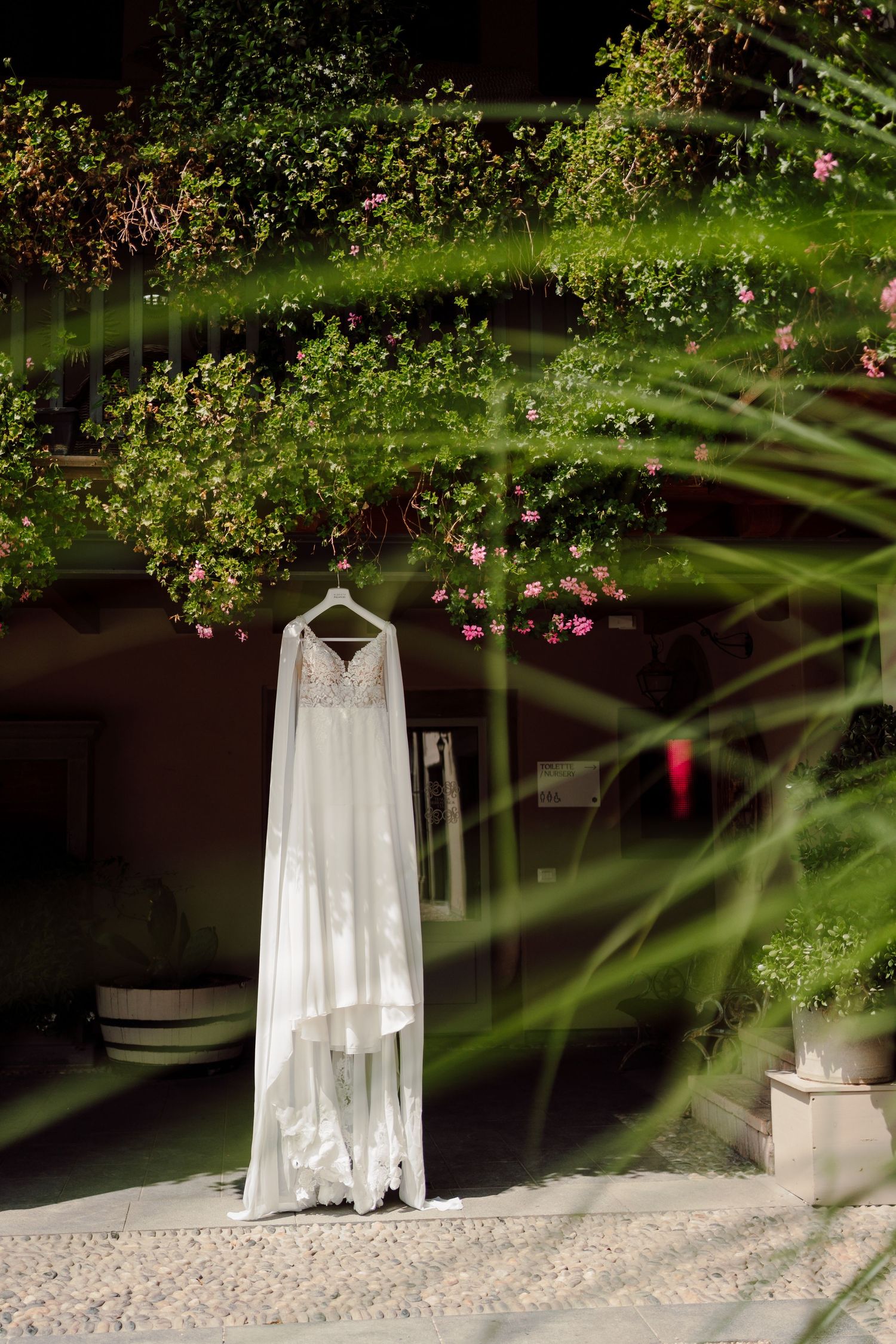 Elegant white wedding dress hanging on a hanger surrounded by lush green tropical plants and flowers.