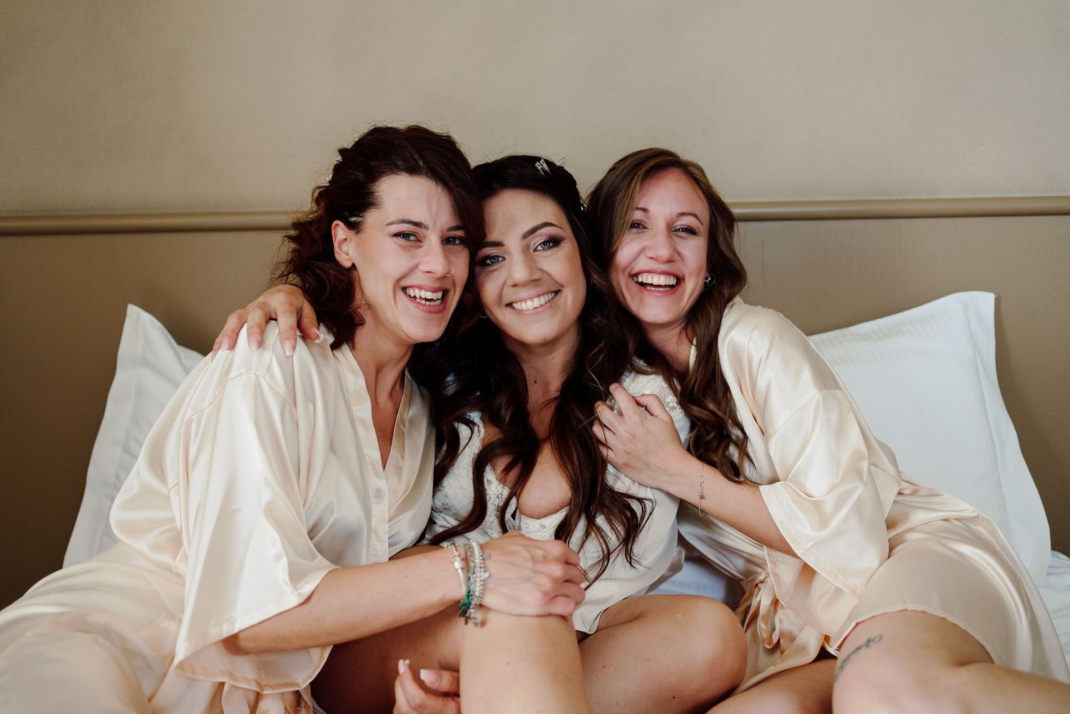 Bride and two bridesmaids in white robes smiling and hugging on a hotel bed.