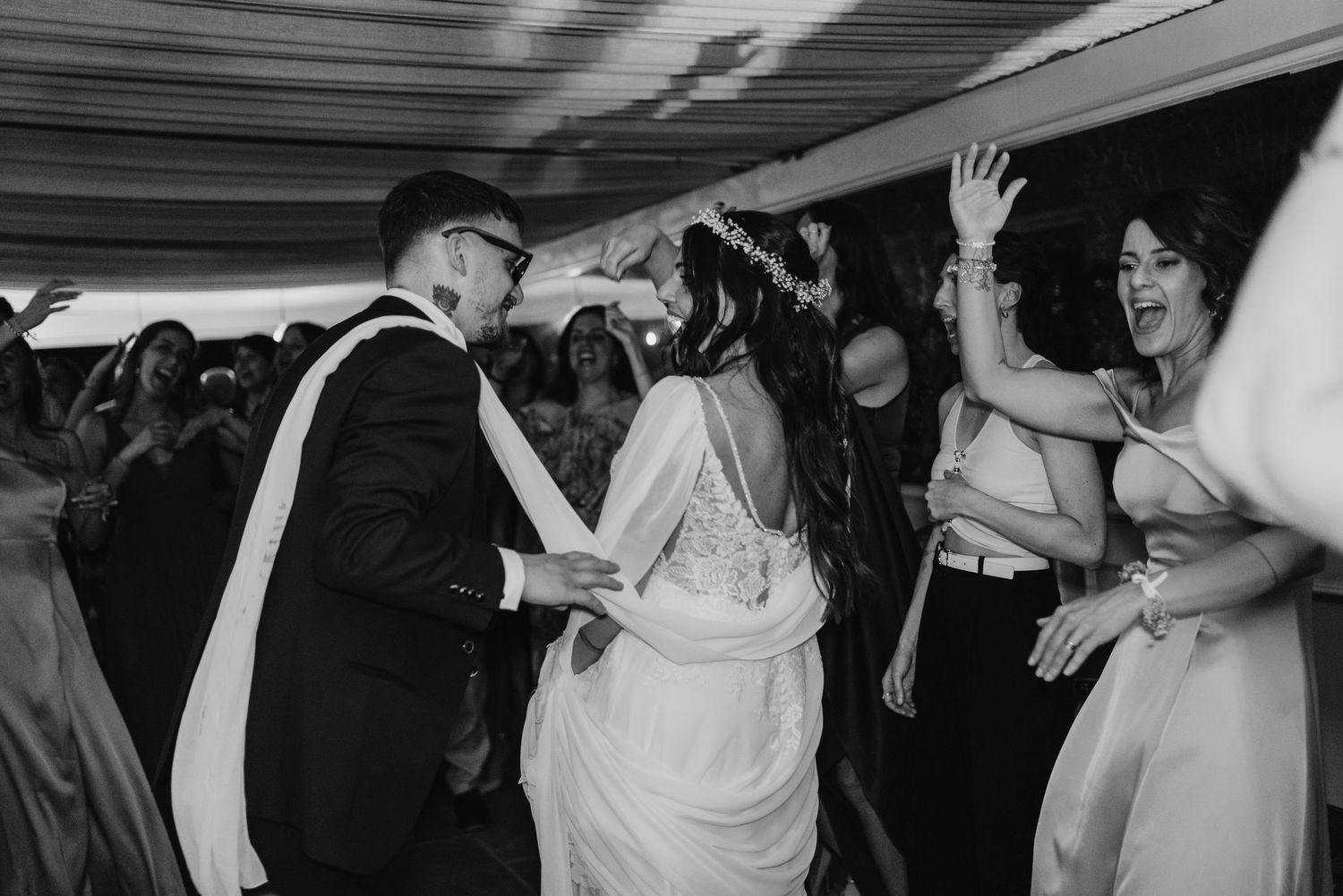 Black and white photo of bride and groom dancing joyfully with guests at lively wedding reception.