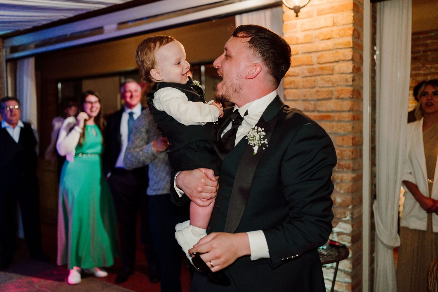 Groom in black tuxedo joyfully lifts smiling child at wedding reception with guests celebrating in background.