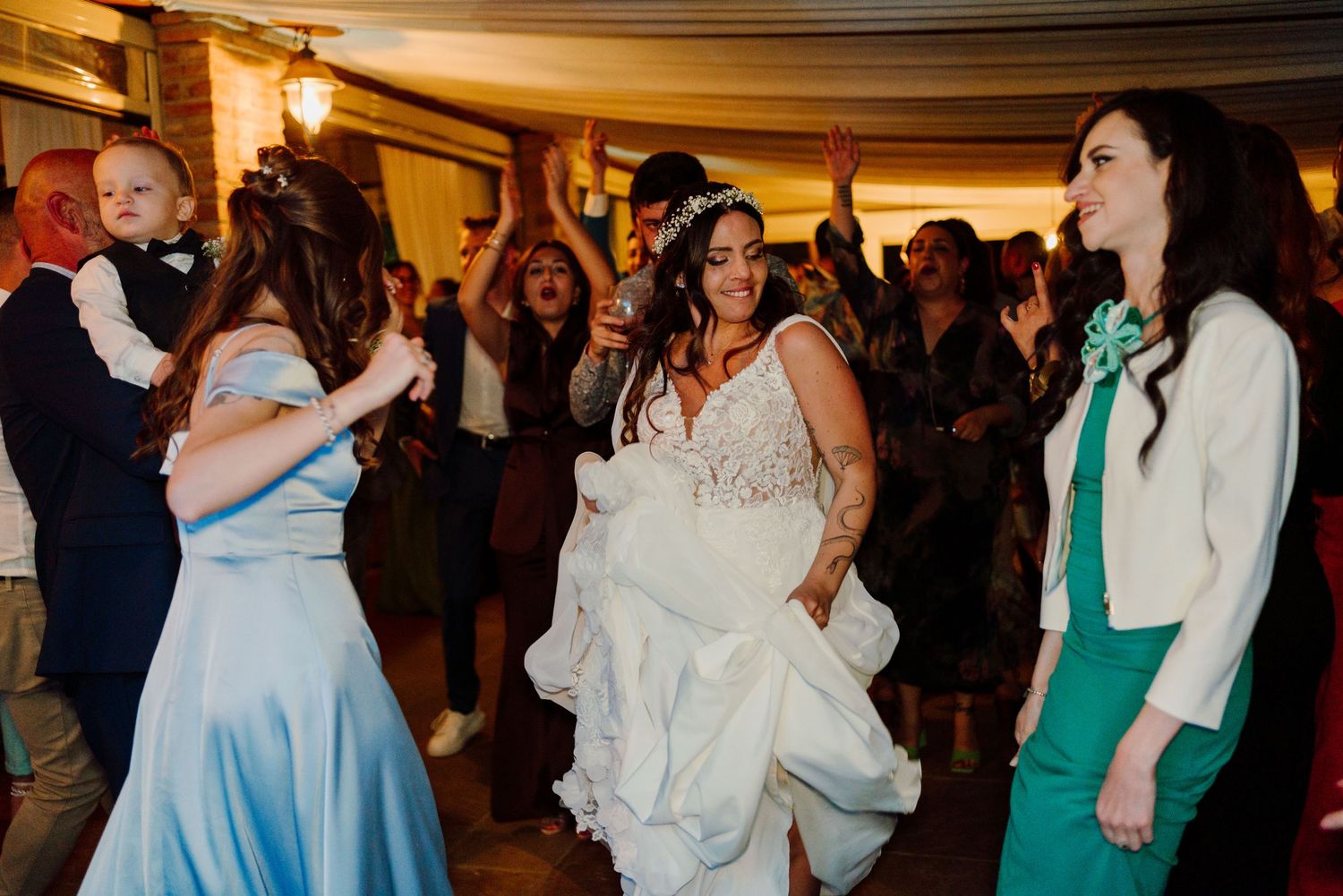 Bride in lace wedding gown dances joyfully with guests in elegant dresses at lively wedding reception.