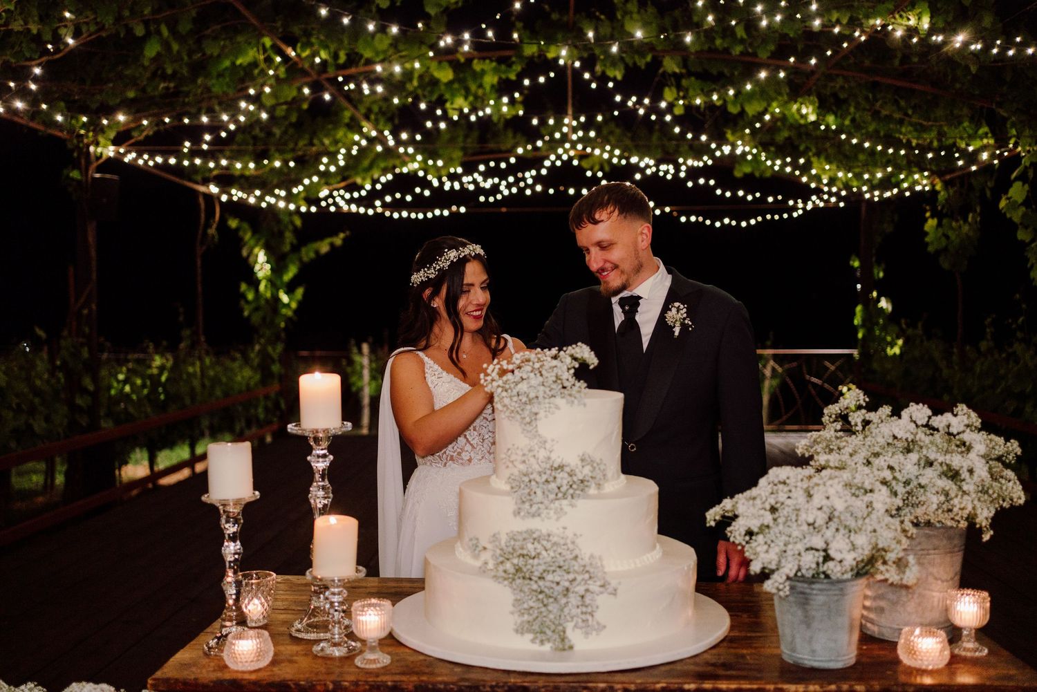 Bride and groom cut elegant four-tier white wedding cake adorned with baby's breath under fairy lights.