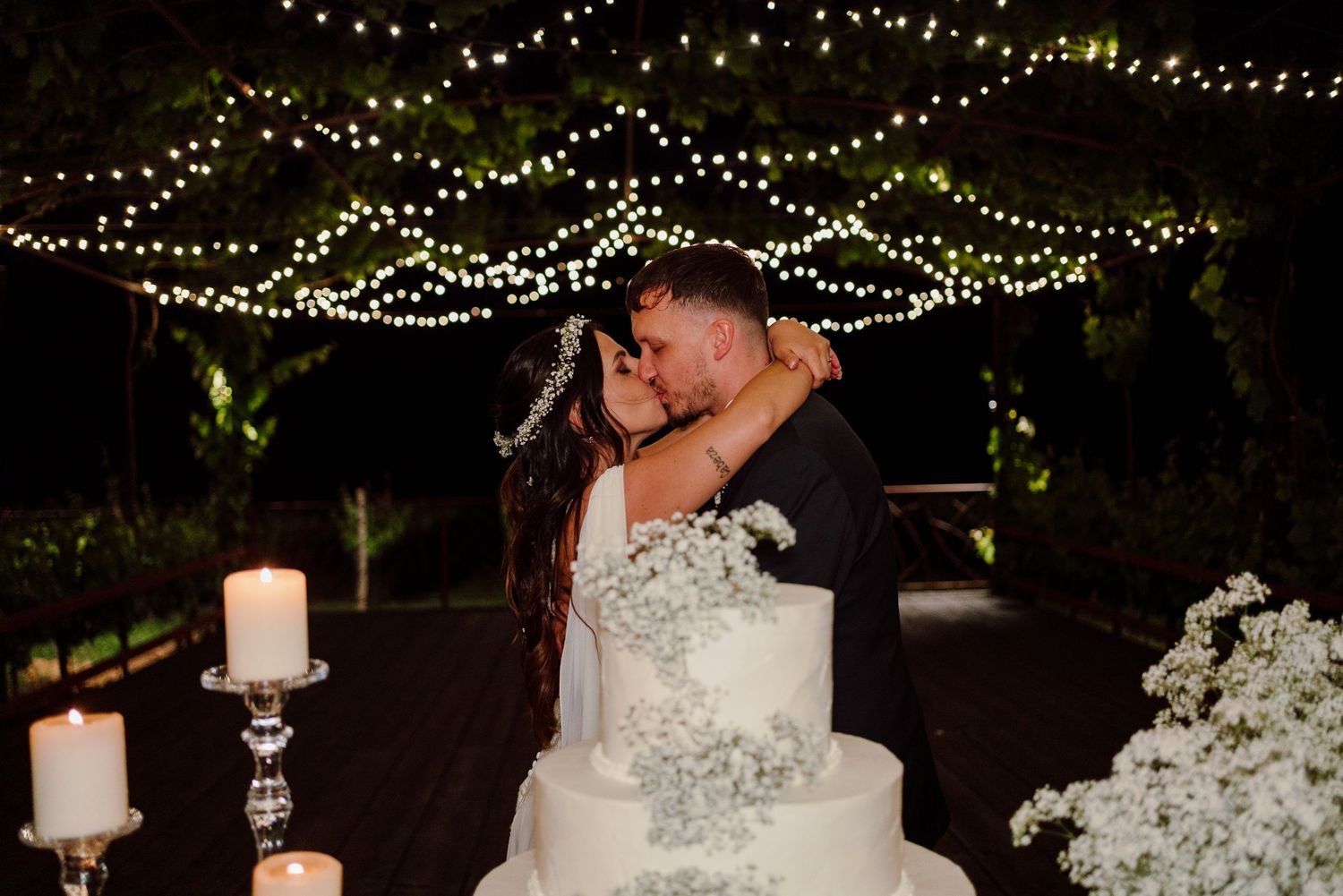 Newlyweds share romantic kiss behind white tiered wedding cake decorated with baby's breath under fairy lights.