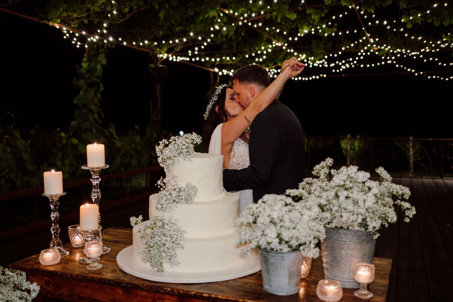 Bride and groom embrace tenderly beside white tiered wedding cake with baby's breath under twinkling fairy lights.