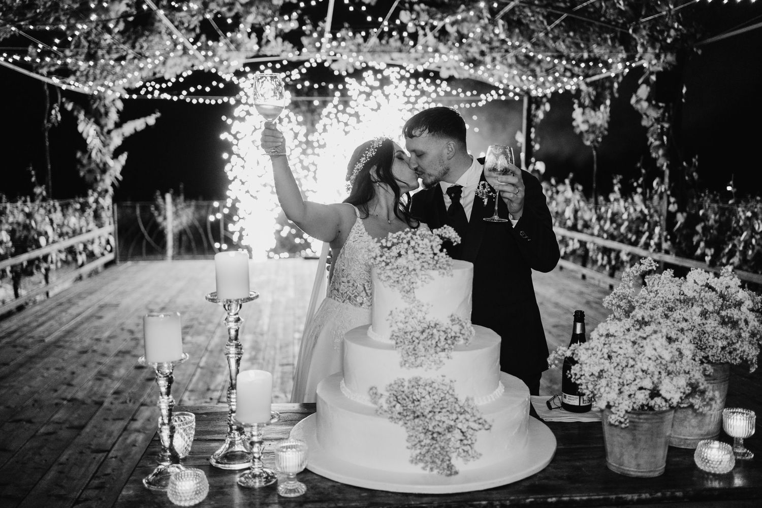 Black and white photo of newlyweds kissing beside tiered wedding cake under sparkling fairy lights at night.