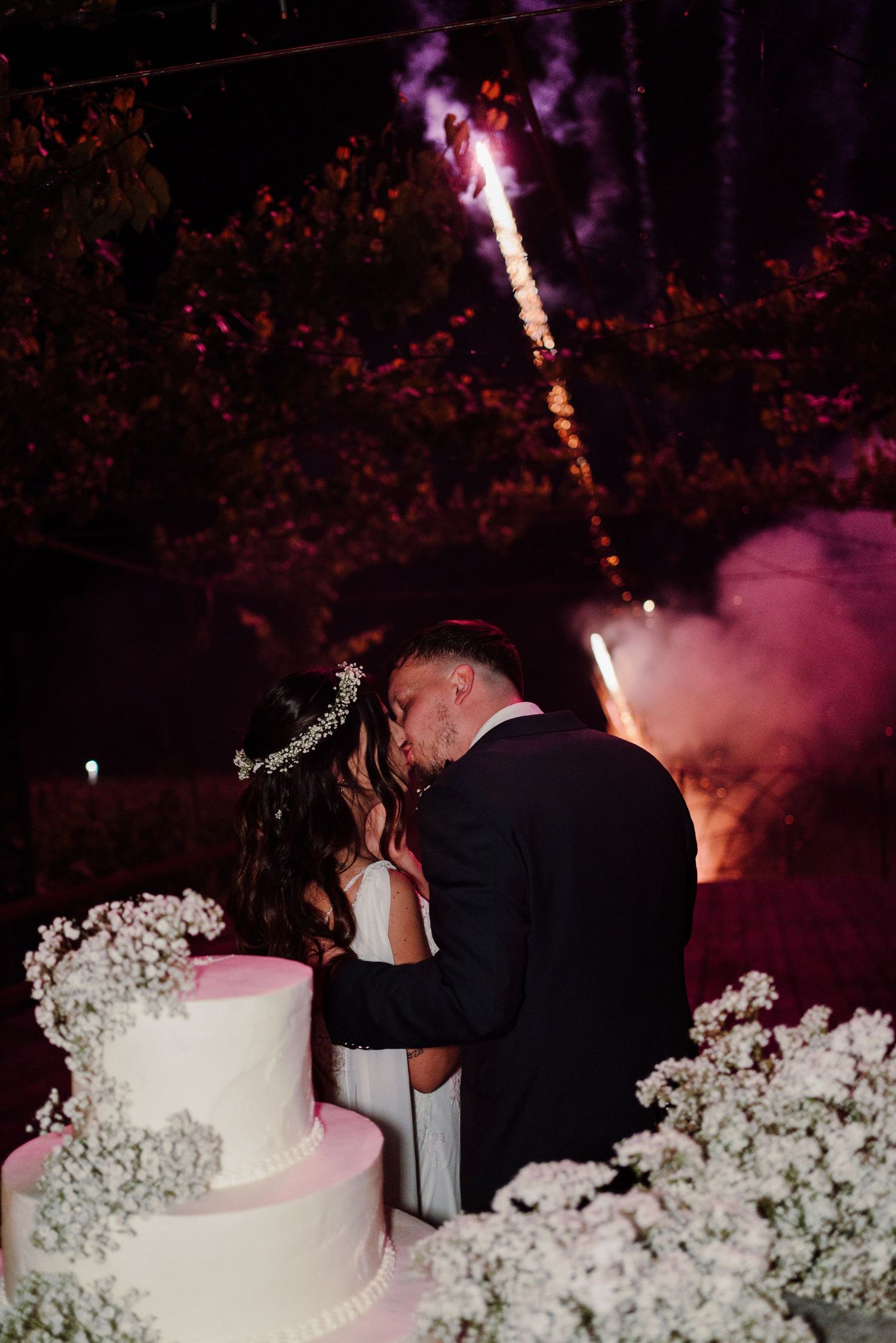 Couple kisses passionately in front of wedding cake with colorful fireworks and smoke in the night background.