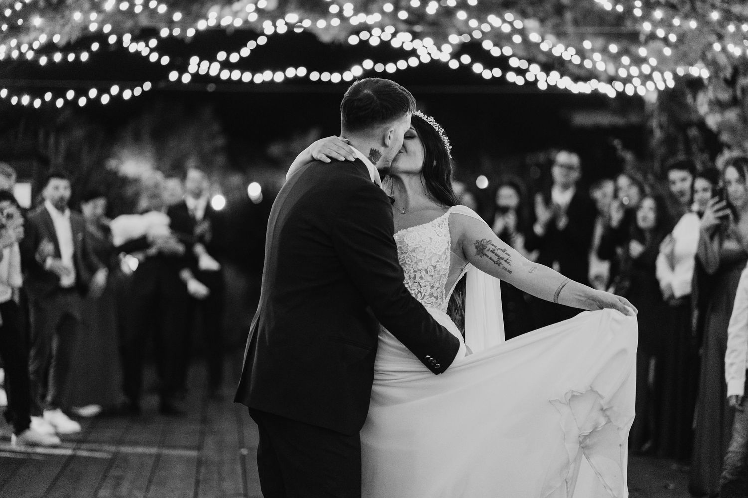 Black and white photo of newlyweds sharing first dance kiss under fairy lights with guests watching.