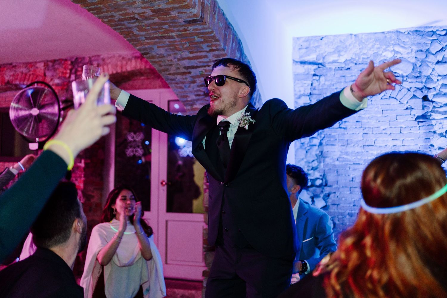 Energetic groom in sunglasses dancing with arms outstretched at lively wedding reception party.