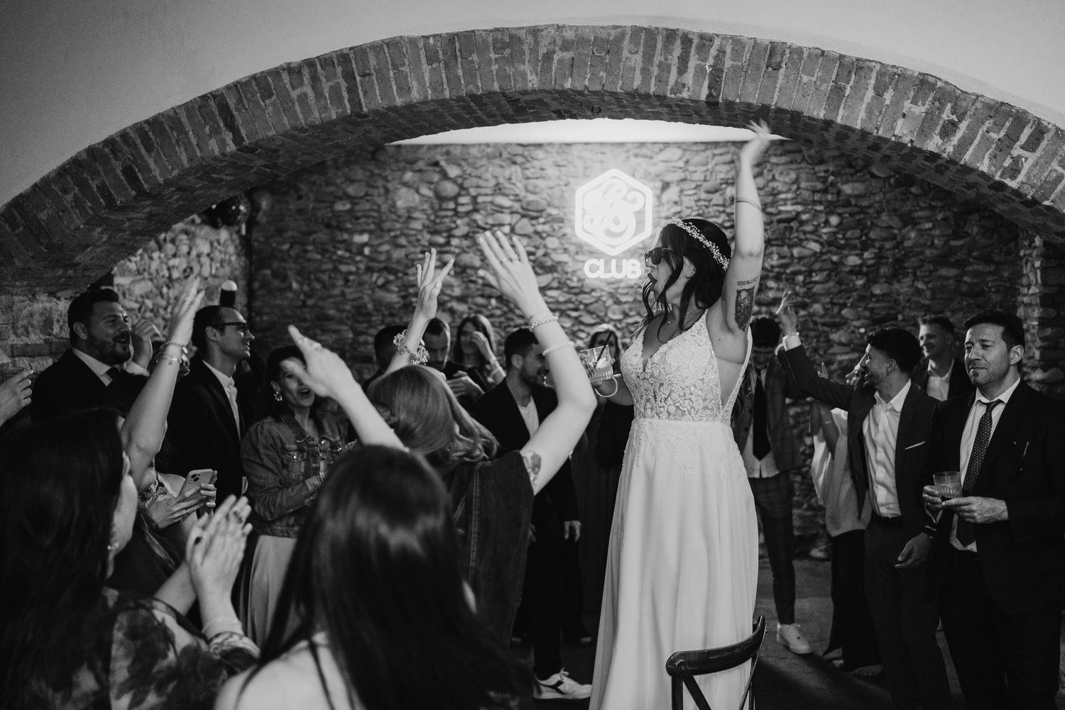 Black and white photo of bride dancing with raised arms under stone arch at wedding reception.