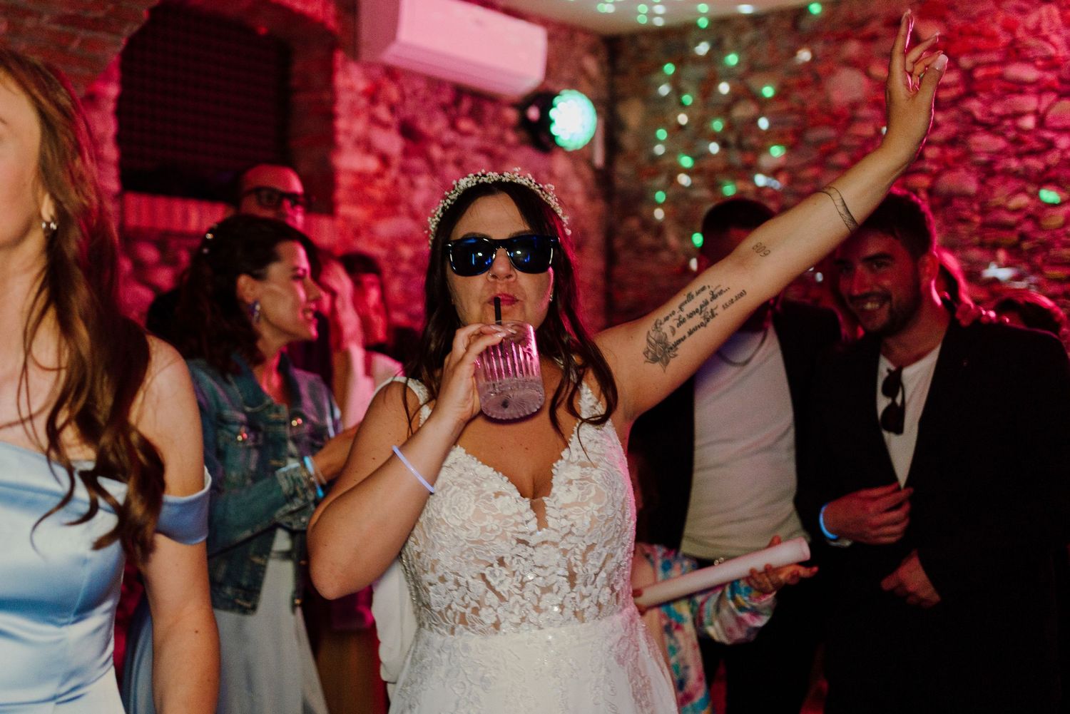 Bride in lace wedding dress wearing sunglasses dancing with guests at vibrant wedding party.