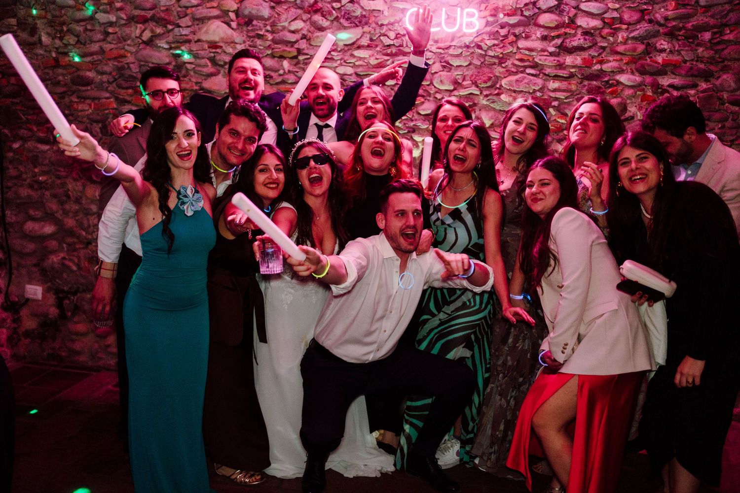 Large group of wedding guests posing and celebrating together in front of neon club sign.