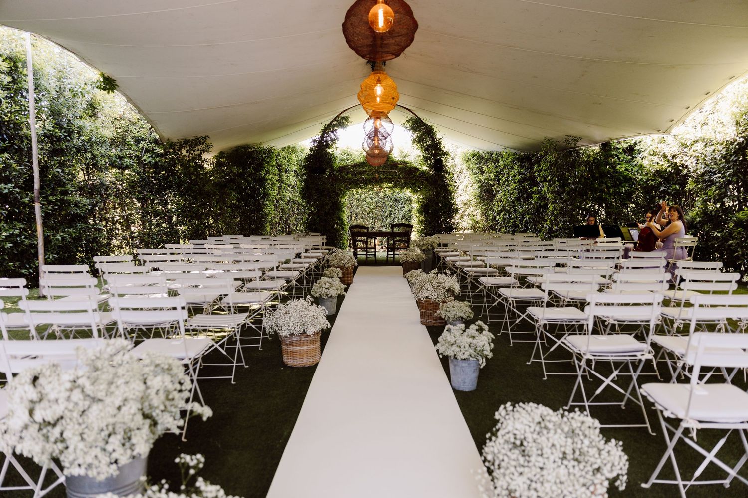 Elegant outdoor wedding ceremony setup with white chairs, baby's breath flowers, and lush green ivy walls under a white tent.