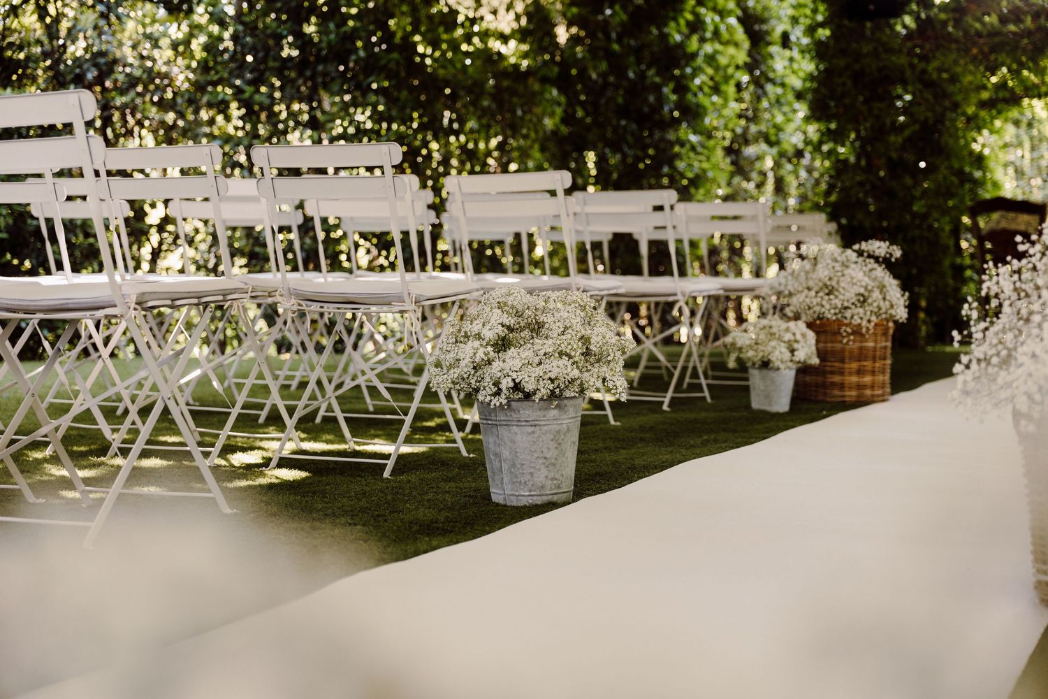 White folding chairs and baby's breath flower arrangements line a white aisle runner at an outdoor garden wedding ceremony.