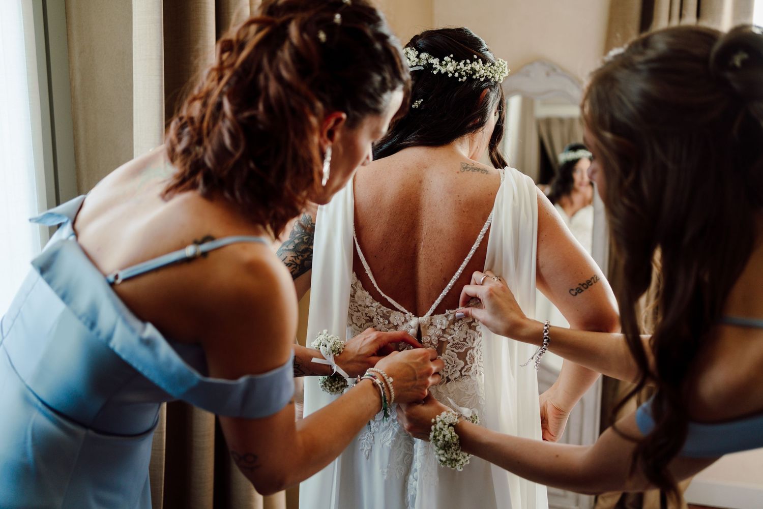 Two bridesmaids in blue dresses helping bride with backless lace wedding gown buttons before the ceremony.
