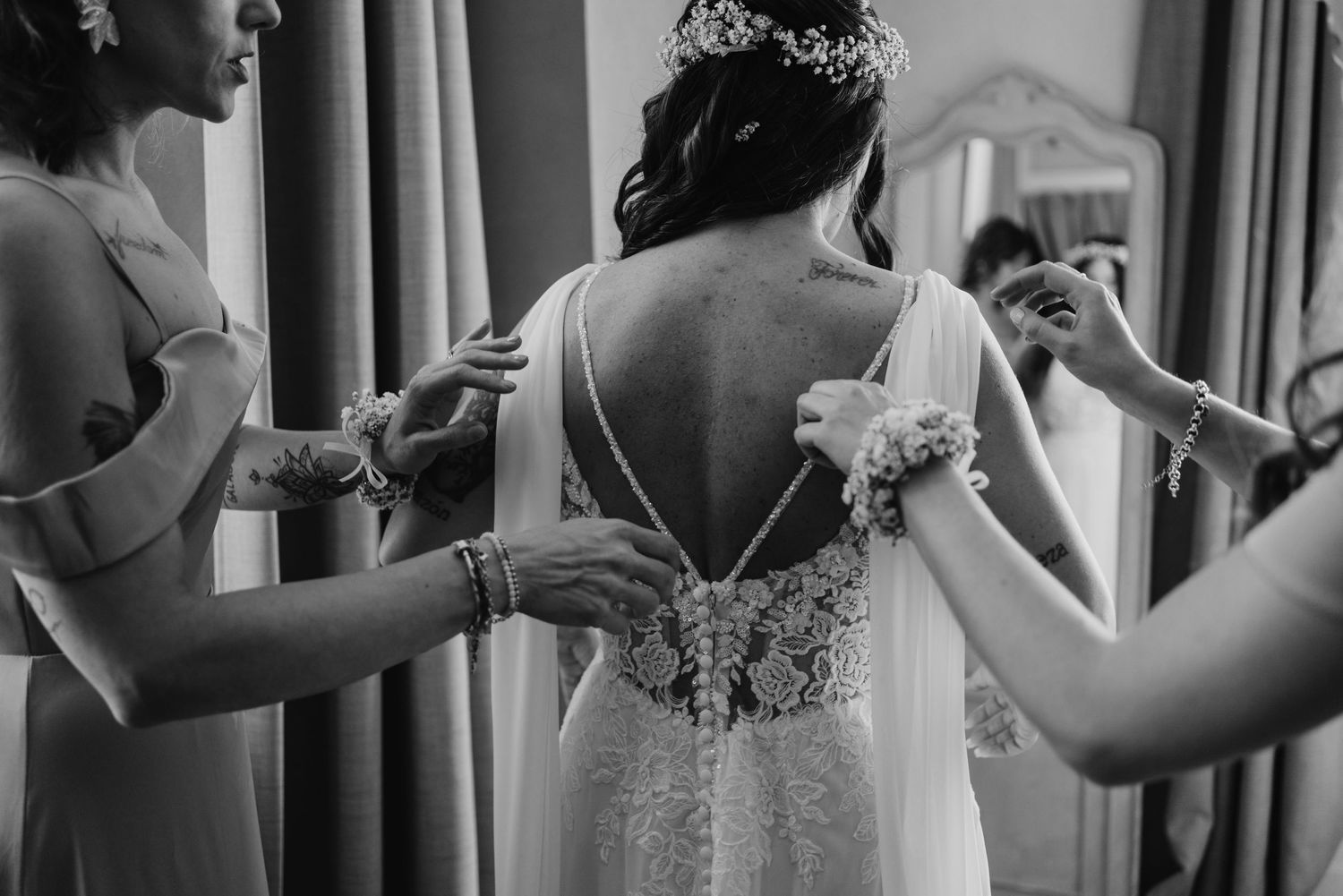 Black and white photo of bridesmaids fastening the back of a bride's elegant lace backless wedding dress.