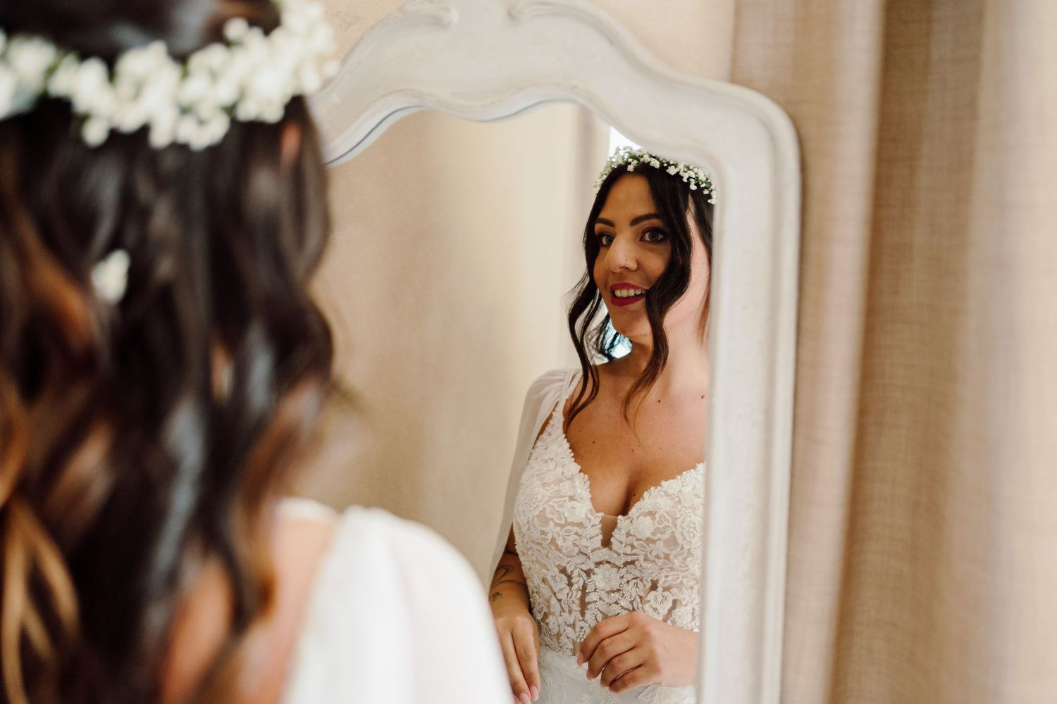 Beautiful bride in lace gown and floral crown admiring her reflection in an ornate white mirror.