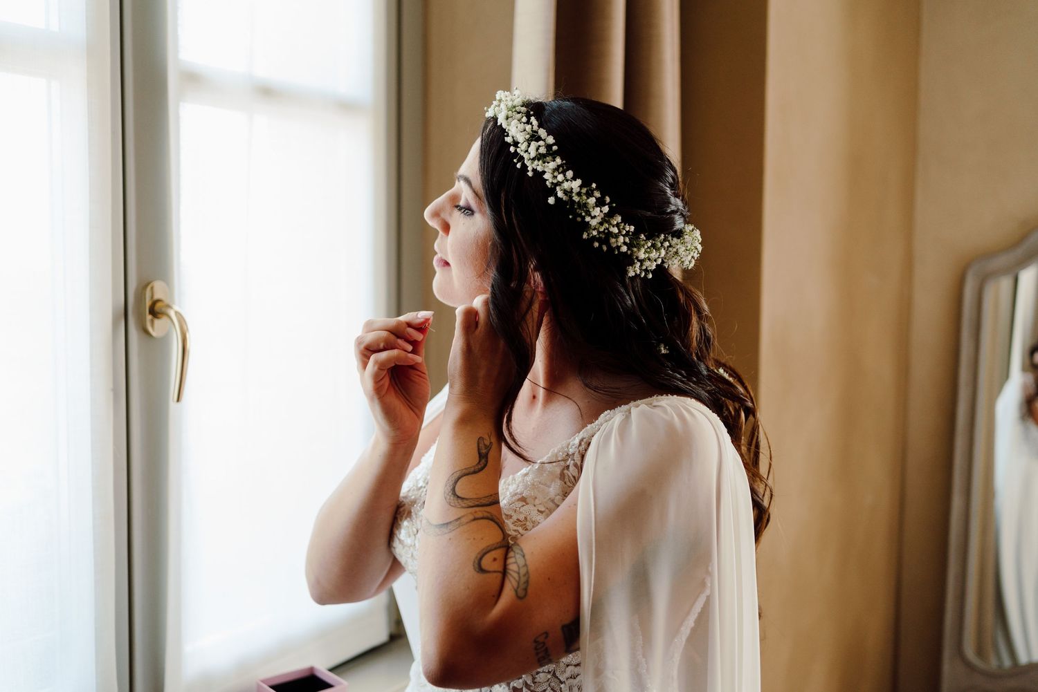Elegant bride wearing a floral crown and lace wedding dress gazing out a window while putting on earrings.