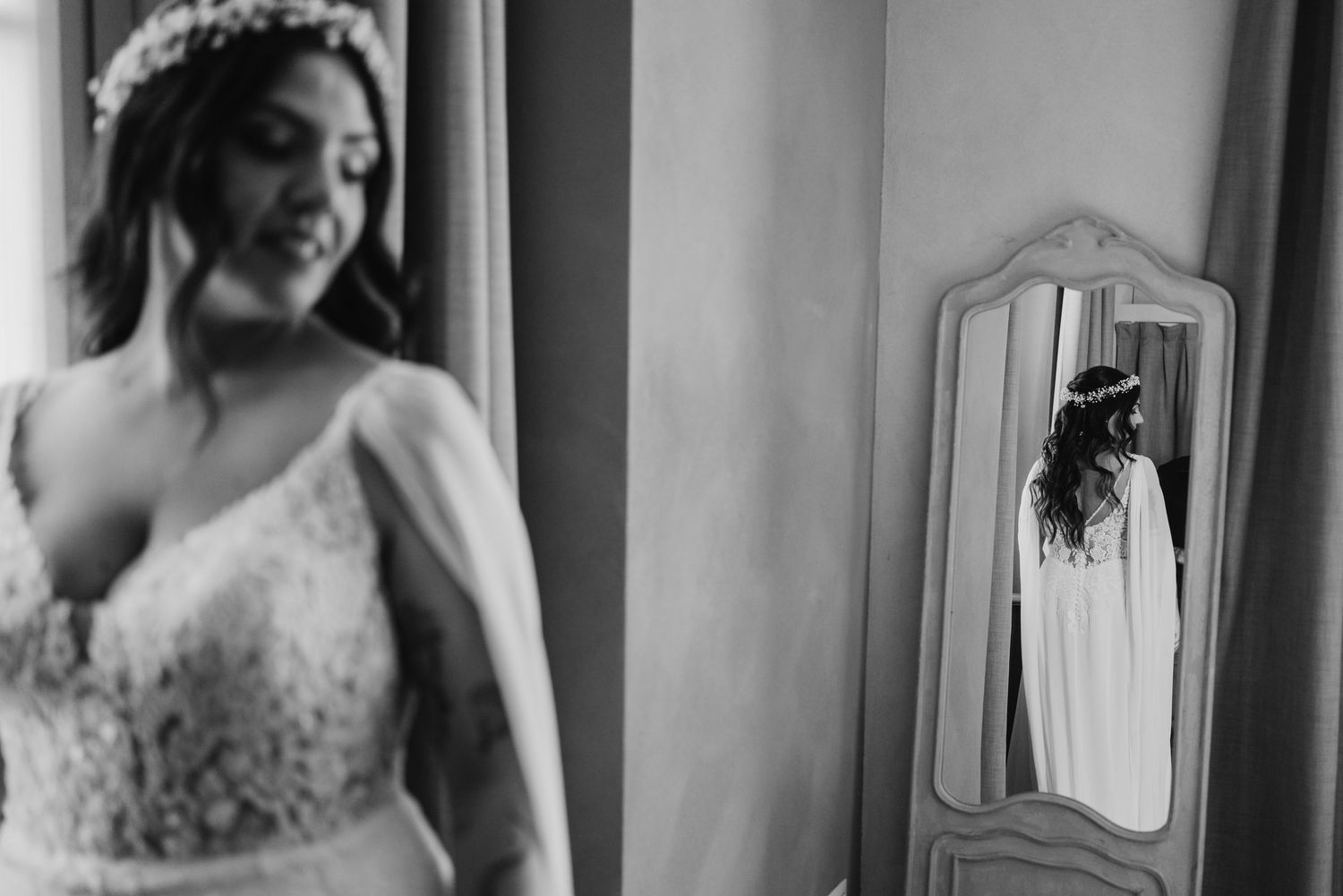 Black and white artistic photo of bride in lace gown with her reflection visible in a tall vintage mirror.