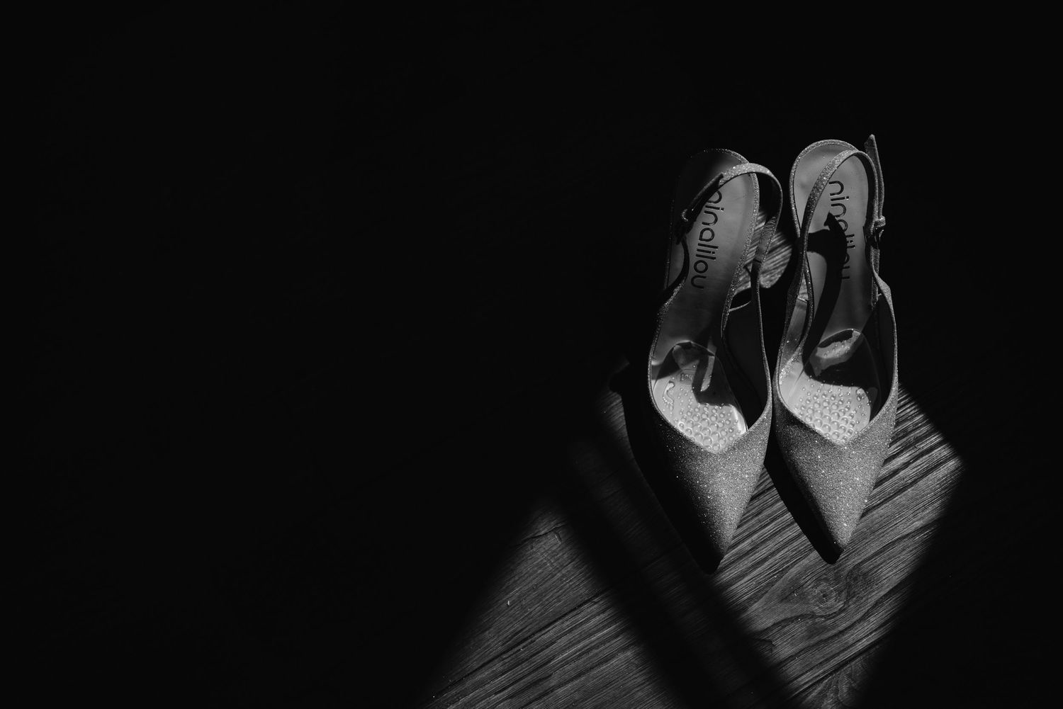 Black and white photo of elegant bridal heels on a wooden floor in dramatic light.