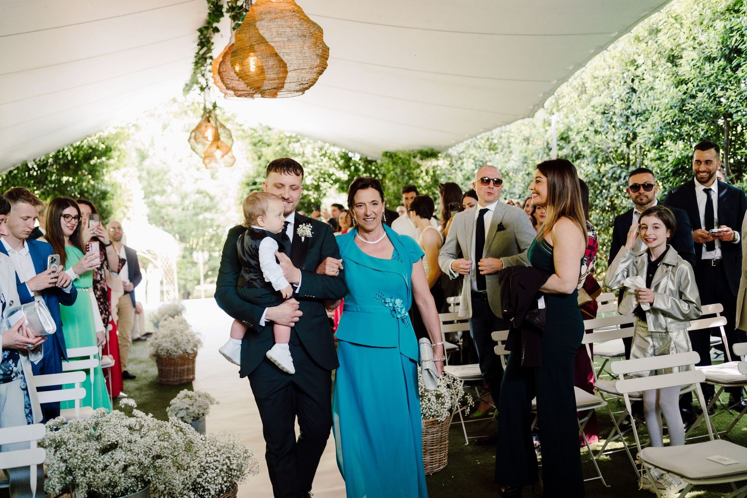Wedding guests walking down the aisle under a white tent decorated with greenery and a Moroccan pendant light.