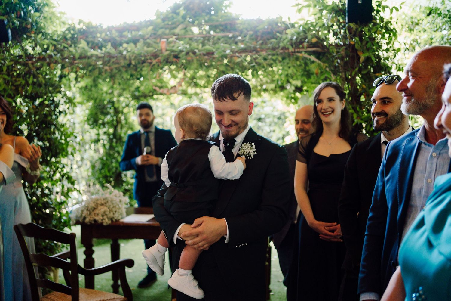 Smiling groom in black suit holding a baby while greeting guests at an outdoor garden wedding ceremony.