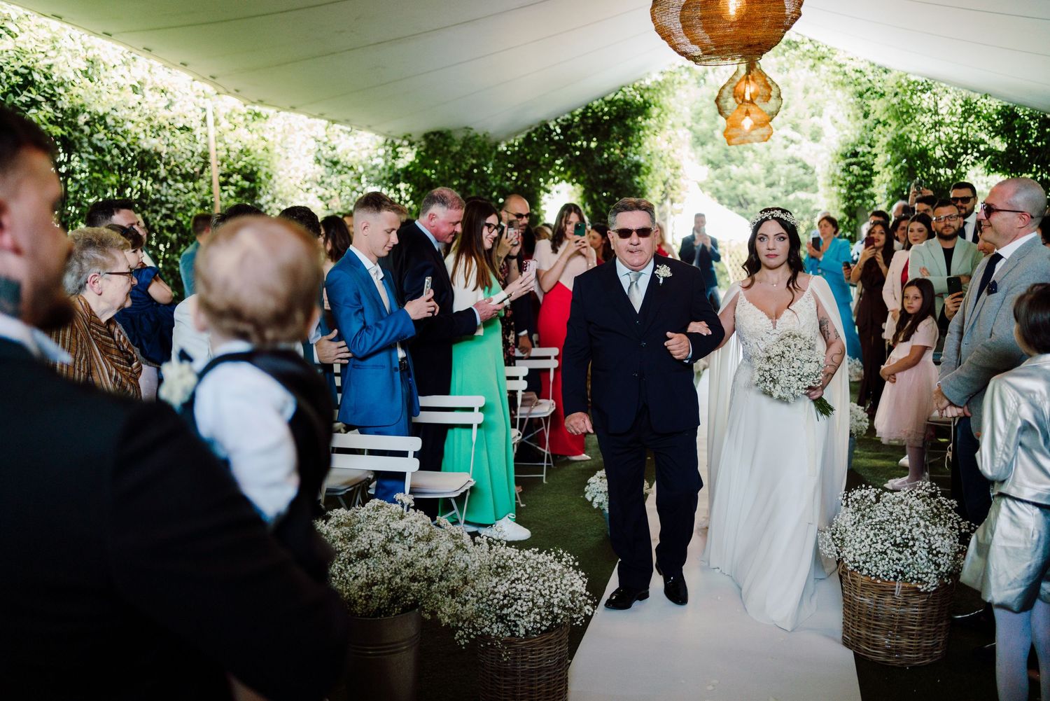 Bride in white lace gown walking down the aisle with her father at an outdoor garden wedding ceremony.