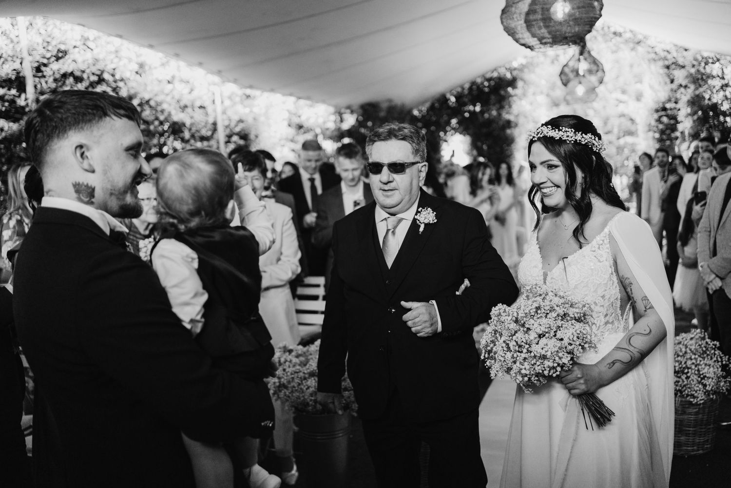 Bride walks down the aisle with her father at an outdoor wedding ceremony under a white tent.