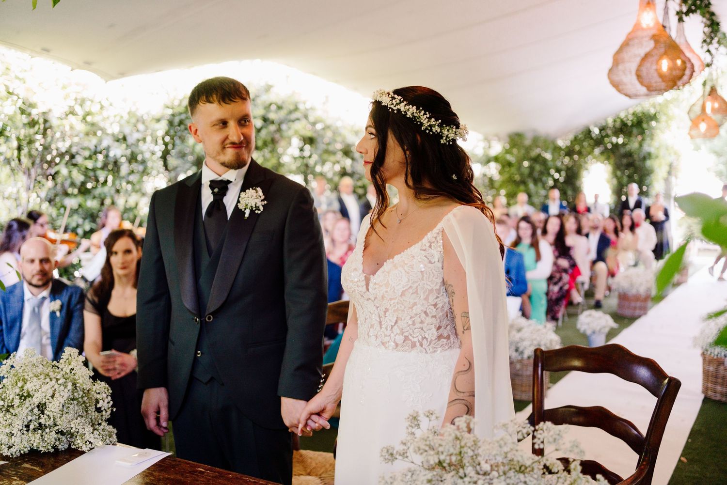 Bride and groom hold hands at the altar during their outdoor wedding ceremony under a floral tent.
