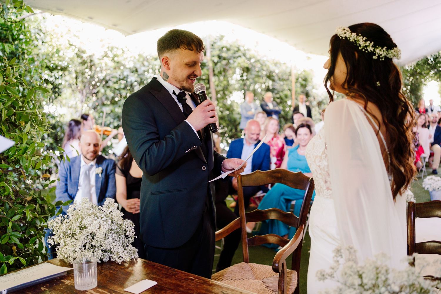 Groom reads wedding vows to bride at rustic outdoor ceremony decorated with baby's breath flowers.