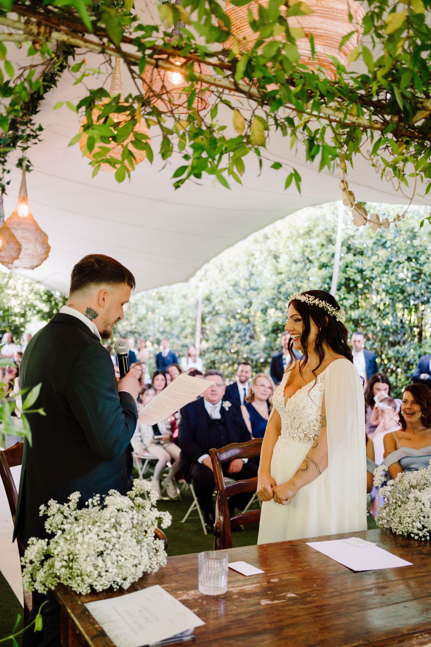 Groom recites vows to smiling bride under a lush green canopy at an outdoor wedding ceremony.