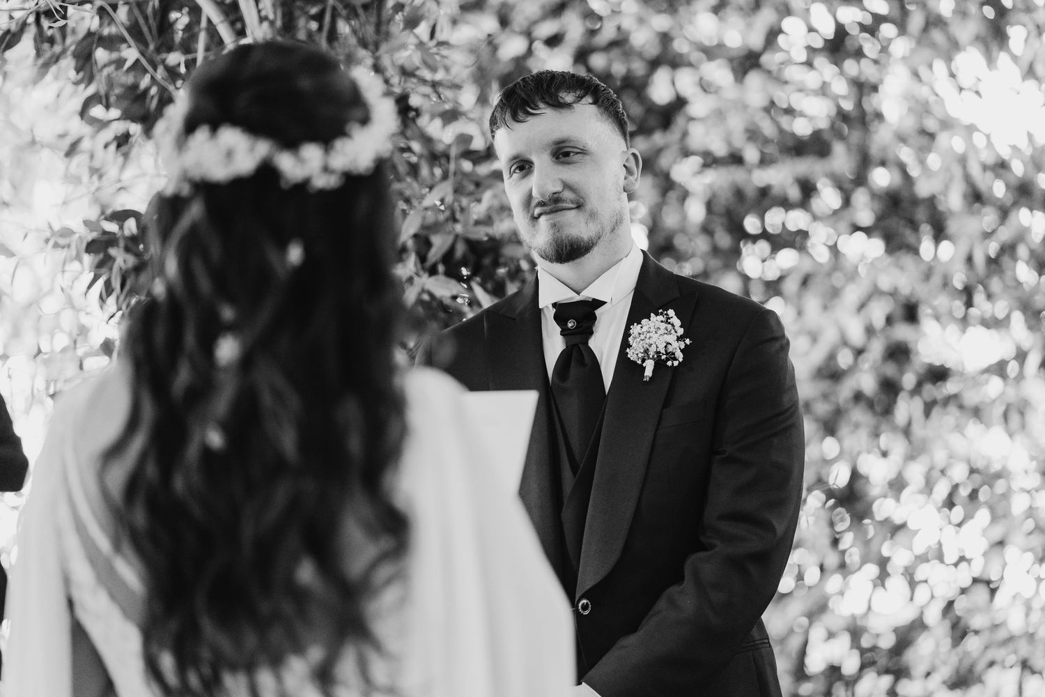 Black and white photo of groom smiling at bride during outdoor wedding ceremony with greenery backdrop.