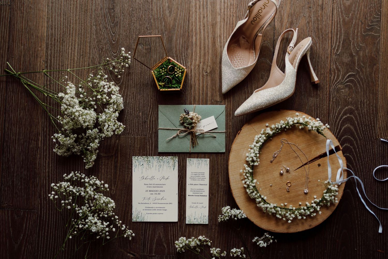 Flat lay of wedding details including invitation suite, floral crown, heels, and greenery.
